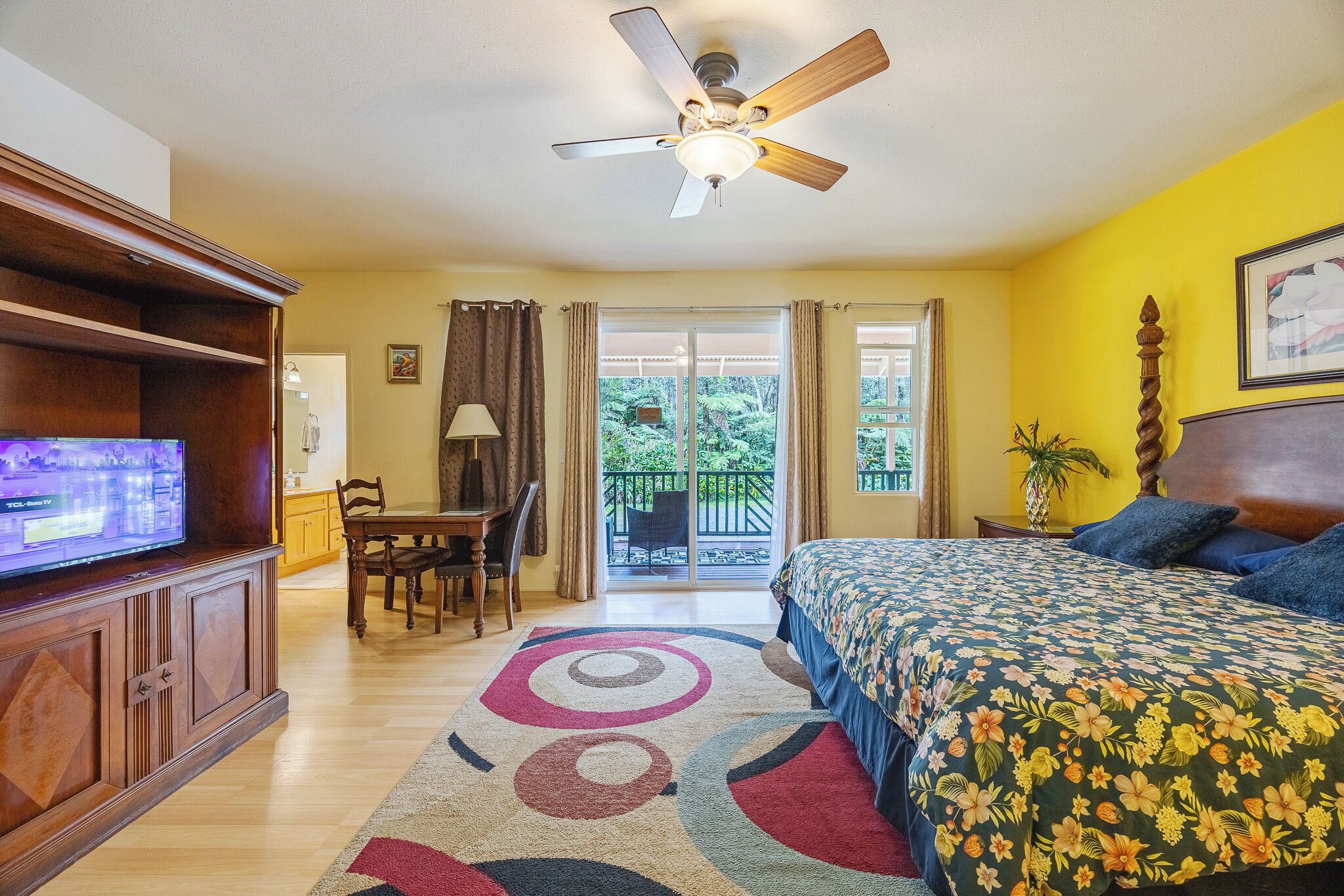 A hotel room with a bed, a TV, a ceiling fan, a dining area, and a balcony with curtains.