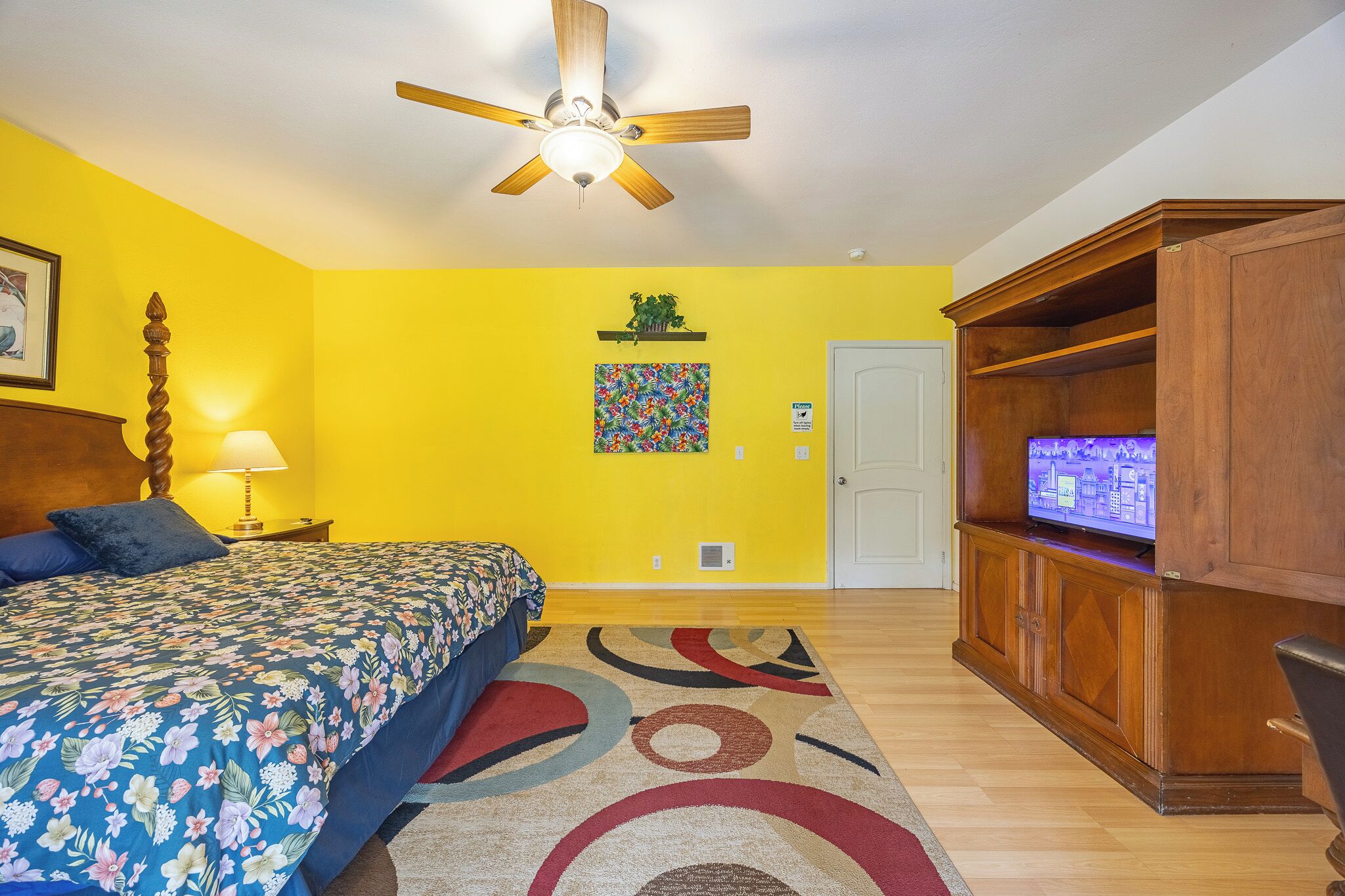 Une pièce avec un lit, un ventilateur de plafond, un téléviseur posé sur une commode en bois et un tapis coloré.