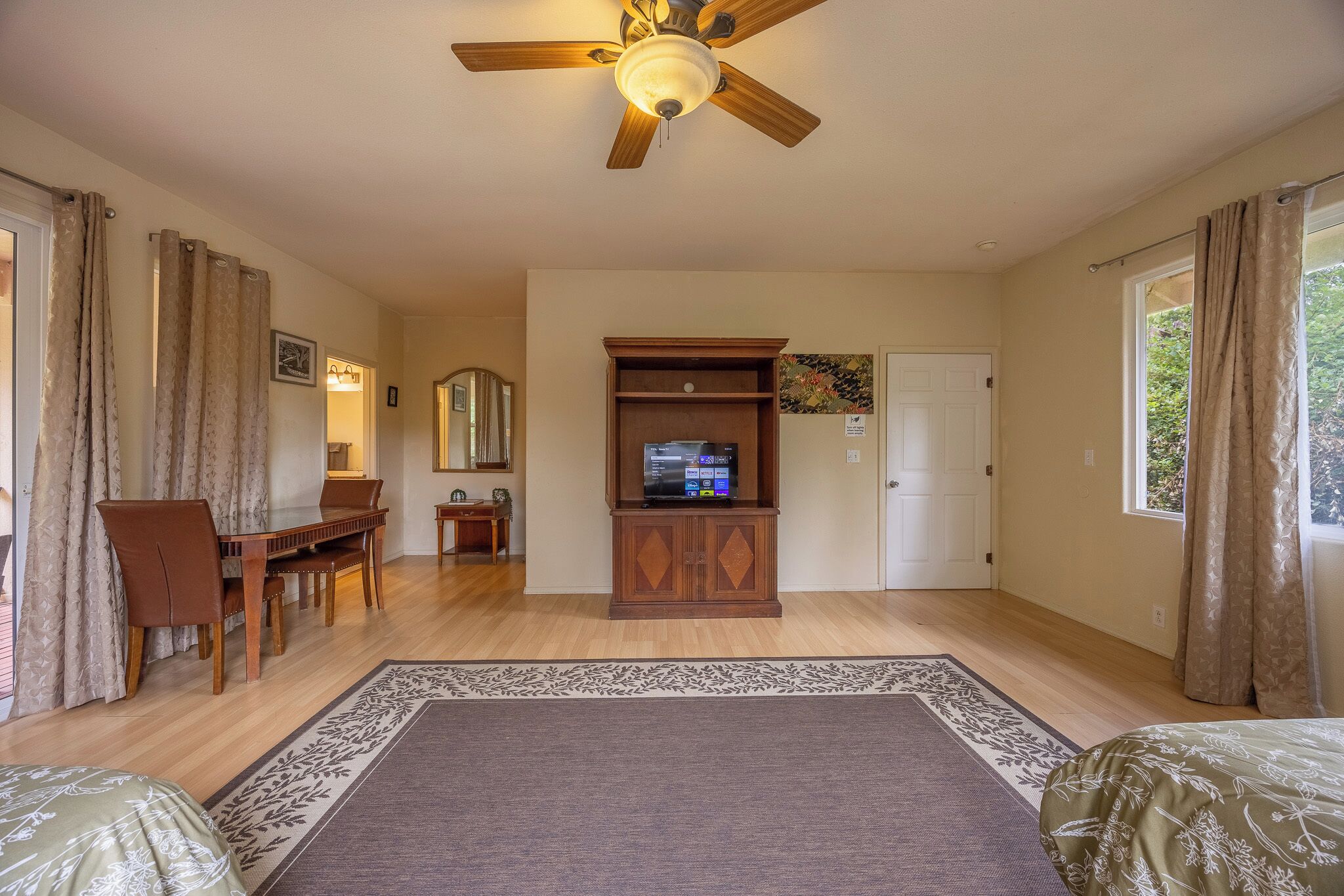 A room with a ceiling fan, a television on a wooden cabinet, a dining table with chairs, and a large window with curtains.