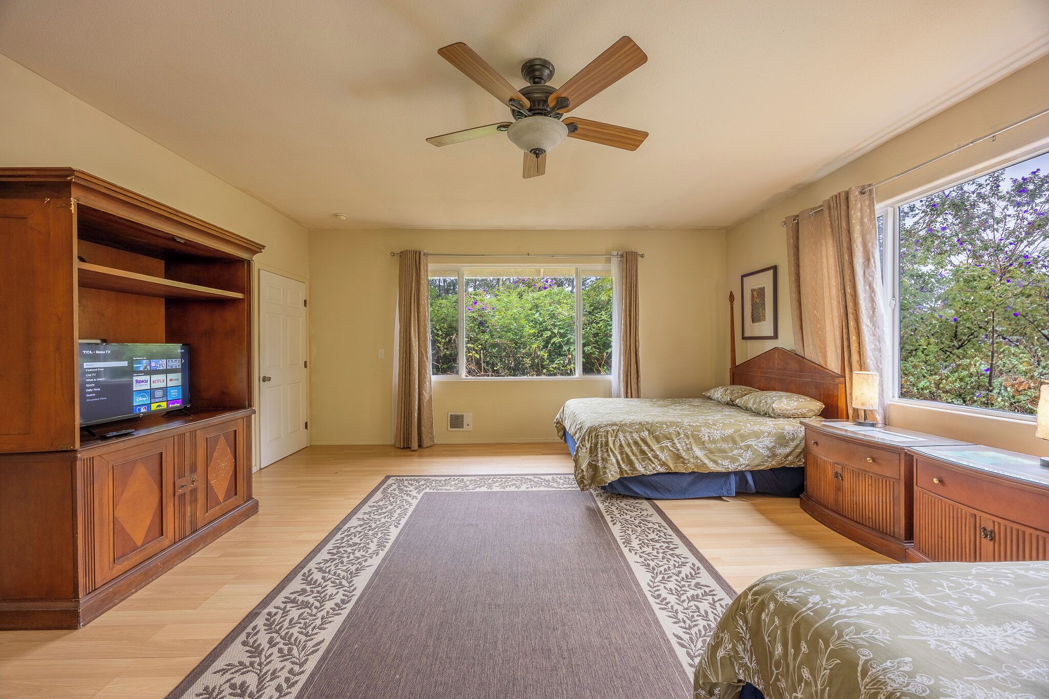 A bedroom with a ceiling fan, a bed with a floral bedspread, a wooden dresser, and a window with a view of trees.