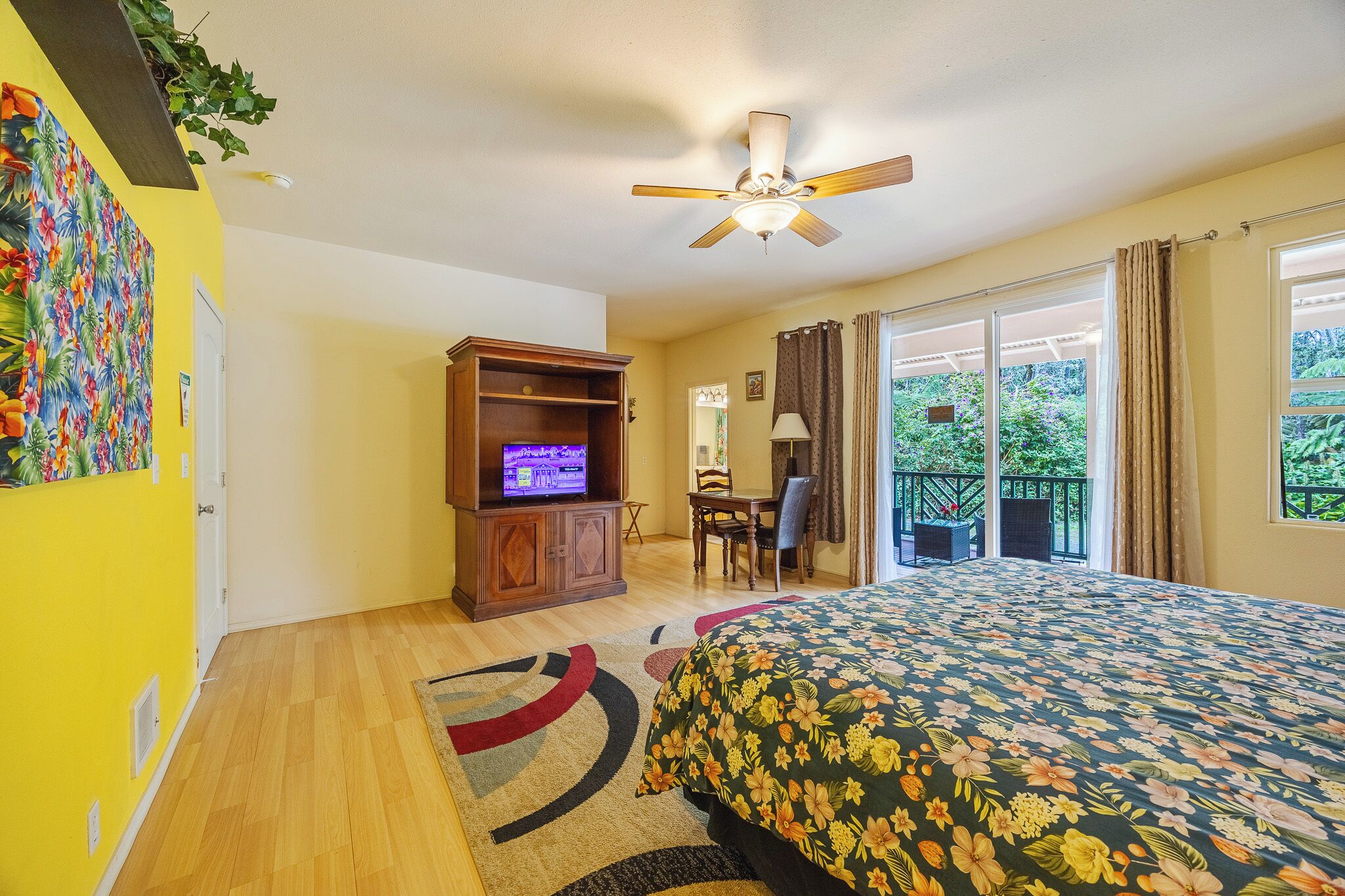 A bedroom with a bed, a TV, a desk, and a balcony with a view of greenery.