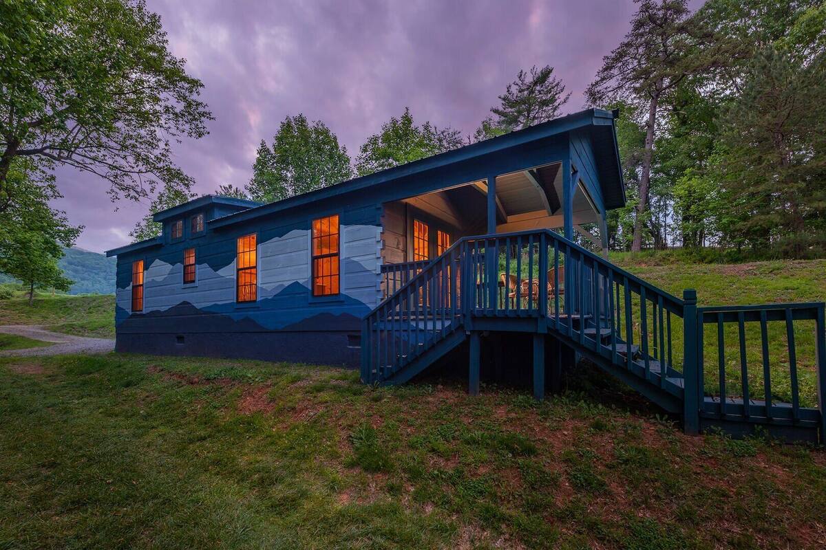 A cabin with a staircase leading to a deck, surrounded by trees and a grassy area.