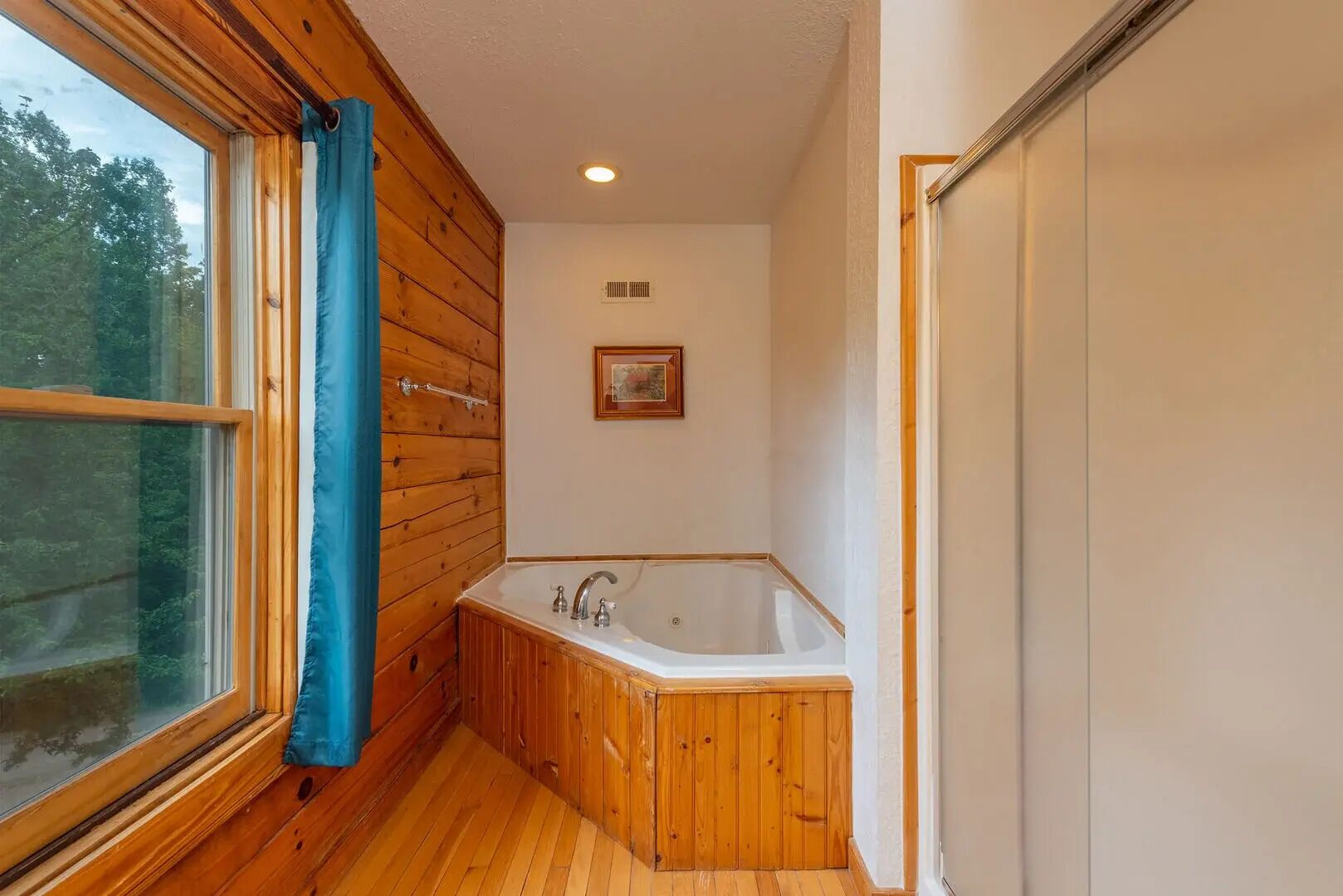 A bathroom with a wooden floor, a bathtub, a window with blue curtains, and a framed picture on the wall.