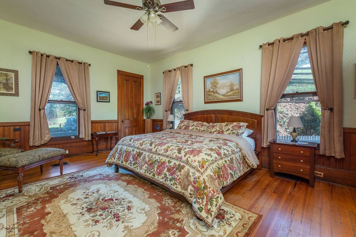 A bedroom with a large bed, wooden floor, a ceiling fan, and multiple windows with curtains.