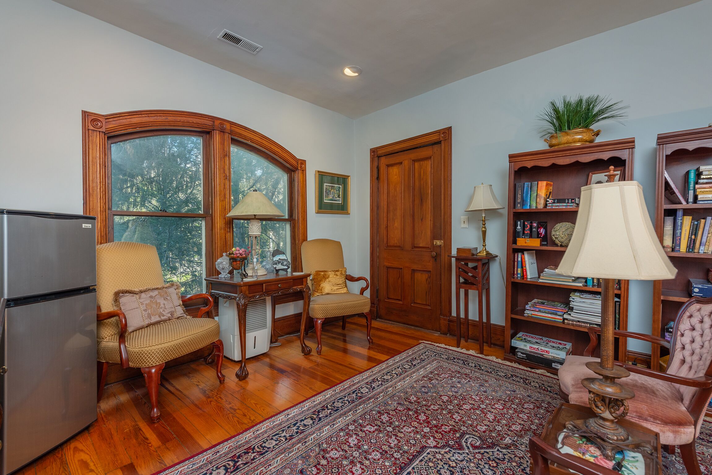 A living room with wooden flooring, a large window, a refrigerator, two armchairs, a rug, a bookshelf, and a door.
