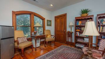 A living room with wooden flooring, a large window, a refrigerator, two armchairs, a rug, a bookshelf, and a door.