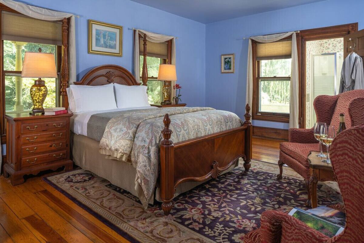 A bedroom with a wooden four-poster bed, two bedside tables with lamps, a chair, and a window with a view of greenery.