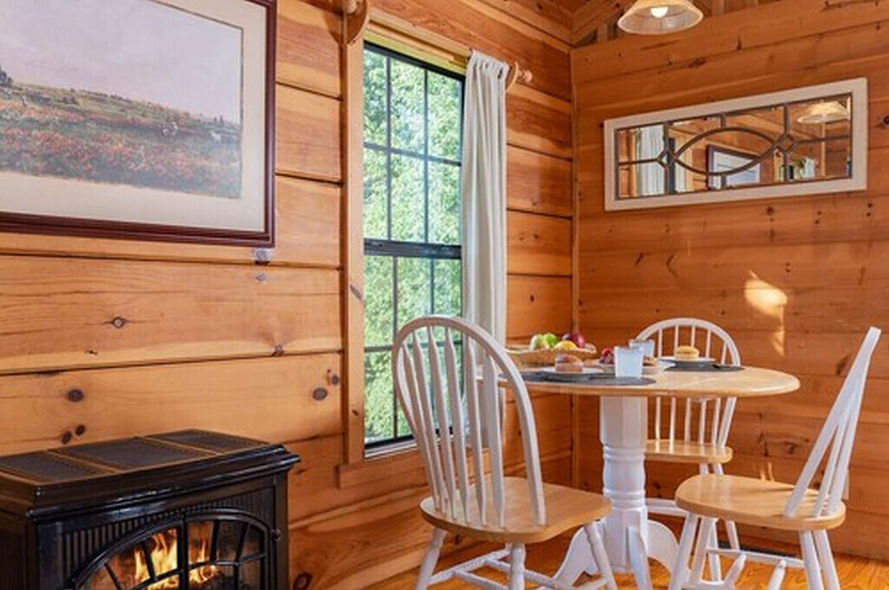 A wooden cabin interior with a dining table, white chairs, a fireplace, and a window with a view of greenery.