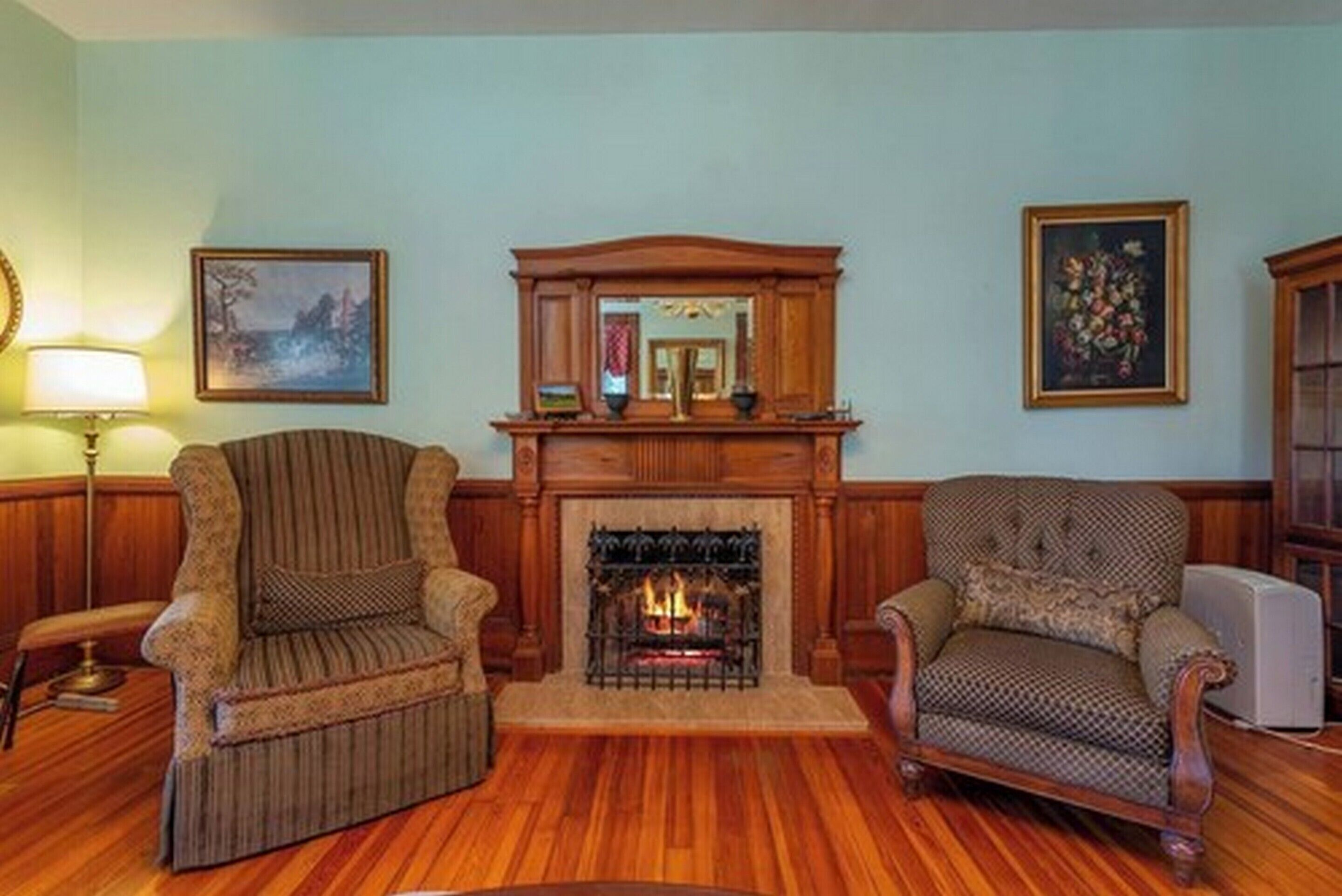 A room with a fireplace, two armchairs, wooden flooring, and framed artwork on the walls.