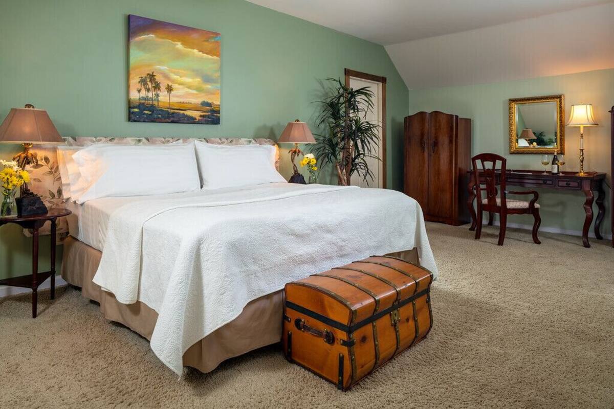 A bedroom with a large bed, a wooden chest, a desk with a mirror, and a painting on the wall.