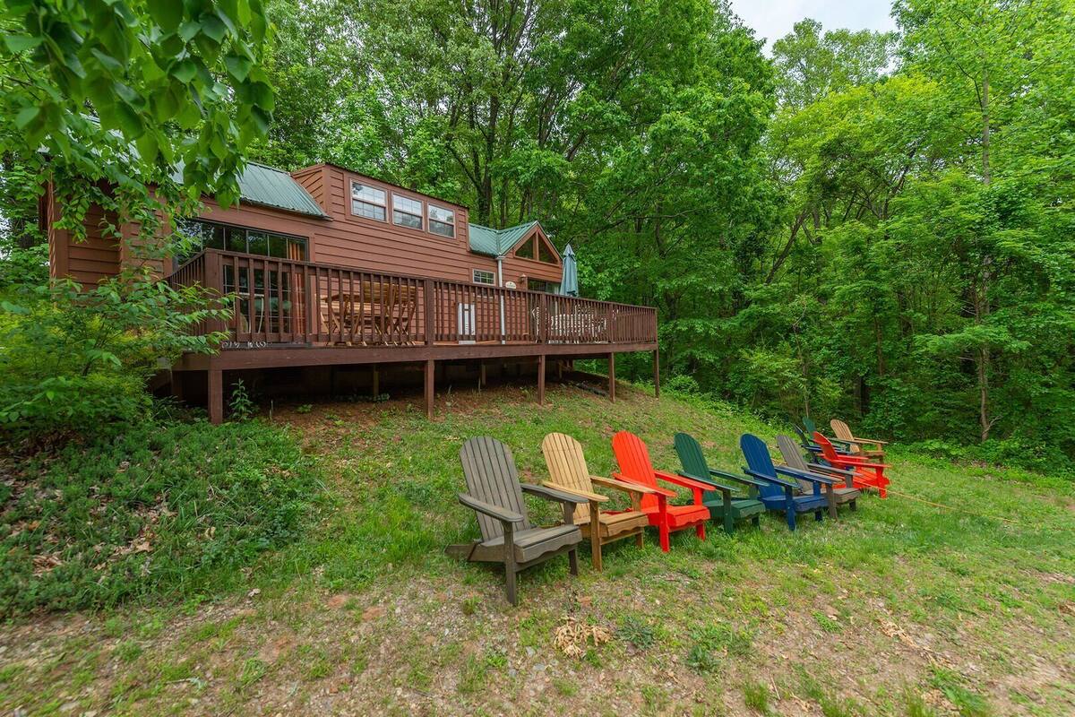 A cabin with a deck, surrounded by a forest. There are several Adirondack chairs on the ground.