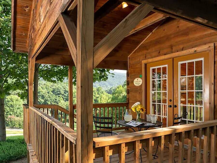Una cabaña de madera con un porche cubierto, una mesa de comedor con sillas y vistas al bosque circundante.