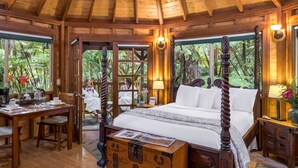 A wooden four-poster bed with a wooden chest of drawers in front, a dining table with chairs, and a view of lush greenery through large windows. - Volcano Village Lodge (Volcano)