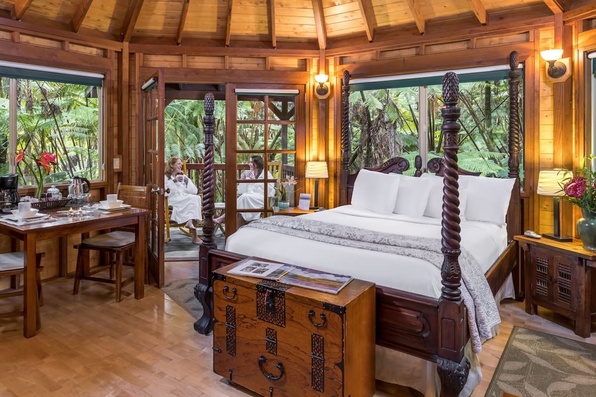 A wooden four-poster bed with a wooden chest of drawers in front, a dining table with chairs, and a view of lush greenery through large windows.
