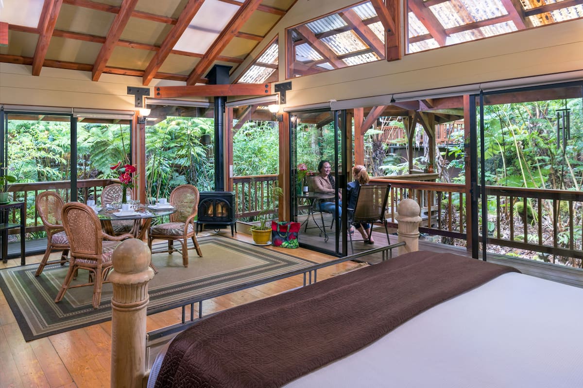 A spacious room with a large bed, a dining area with wicker chairs, and a view of lush greenery through large windows.