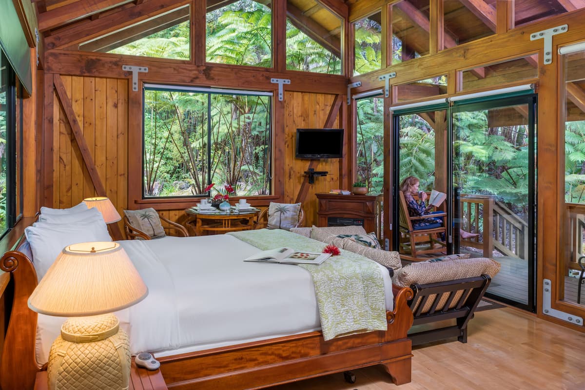 A wooden bedroom with a bed, bedside lamps, a TV, and a view of the forest through large windows.