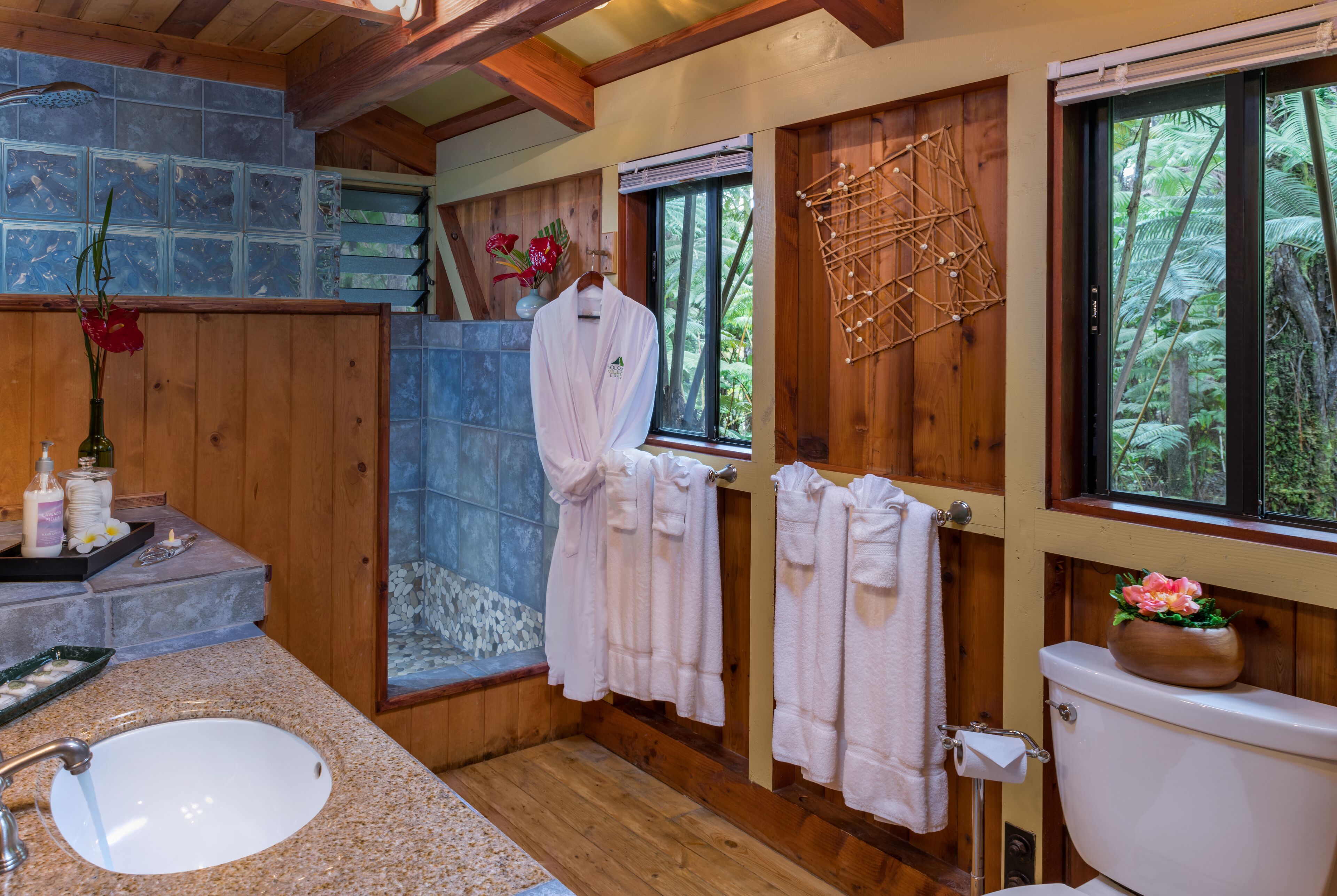Un baño con detalles de madera, un lavamanos blanco y un inodoro. Hay toallas colgadas en un tendedero y una ventana con vistas a árboles.