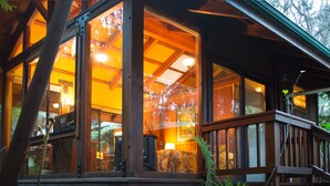 A wooden cabin with large windows, a deck, and a visible interior with a lamp and a patterned rug. - Volcano Village Lodge (Volcano)