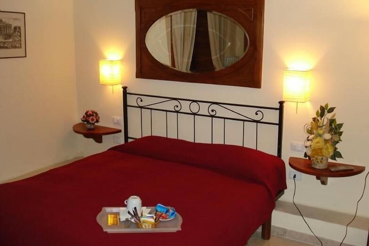 Uma cama bem arrumada com colcha vermelha, uma mesa de cabeceira com um vaso de flores e um espelho fixado na parede.