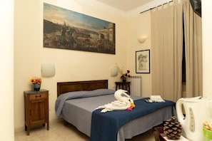 Une chambre d’hôtel avec un lit, un tableau au mur, une petite table avec un vase de fleurs, une bouilloire et une pomme de pin.