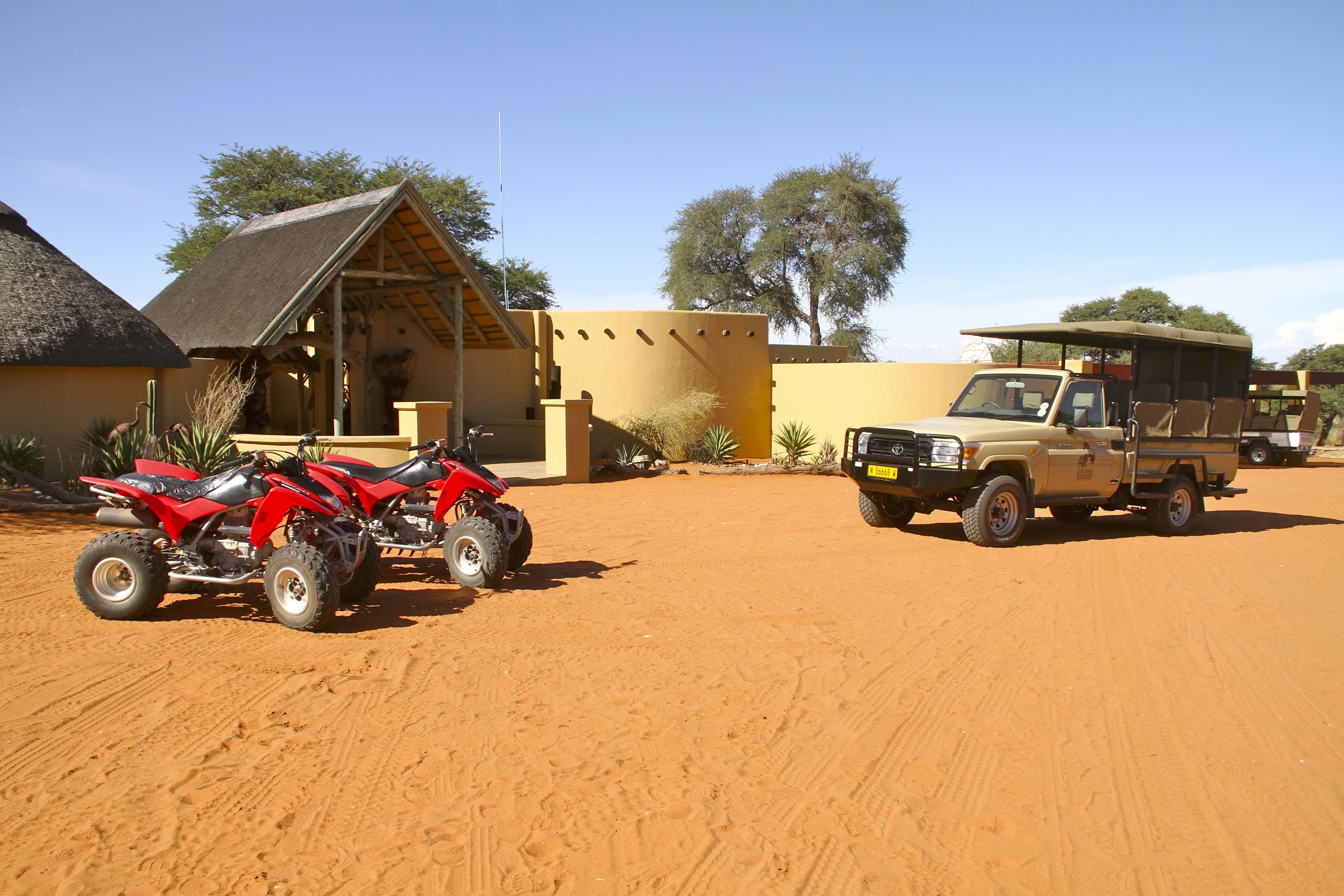 A safari lodge with a thatched-roof building, a beige SUV, and two red off-road vehicles parked on sandy ground.