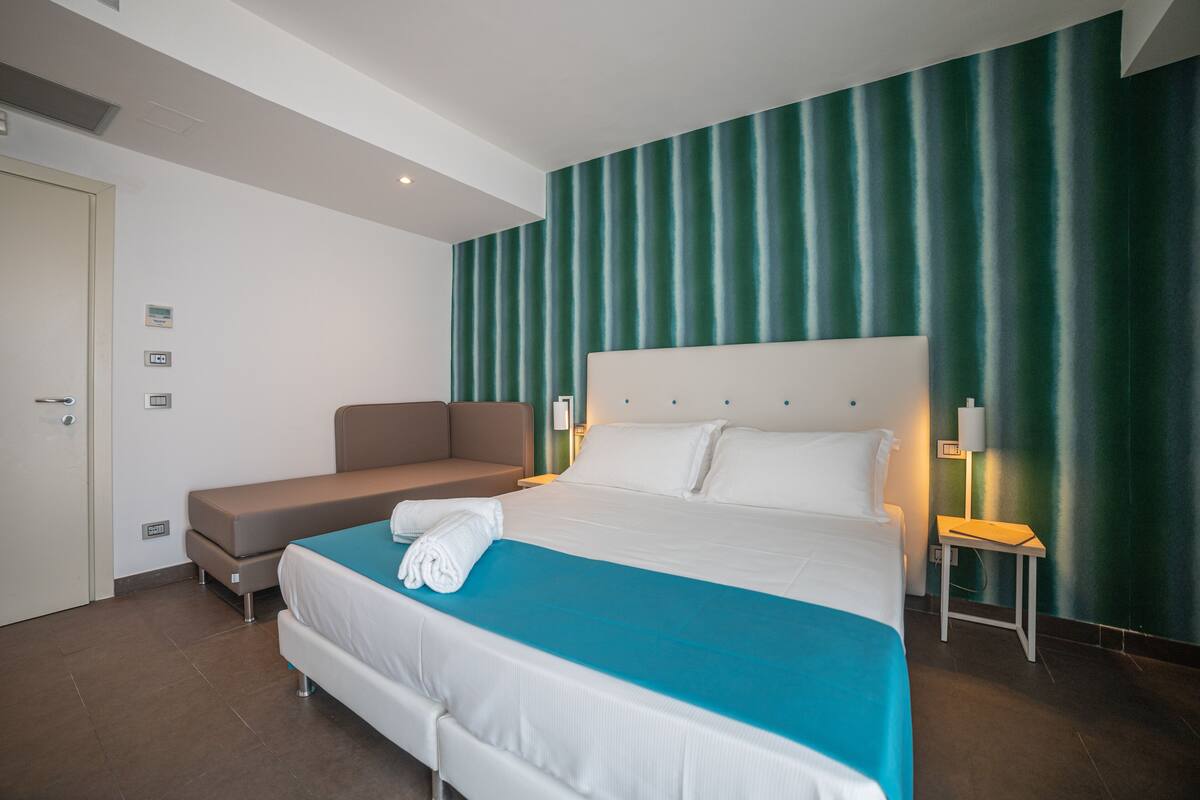 Une chambre d’hôtel avec un grand lit, un canapé-lit, une table de chevet et une lampe fixée au mur.