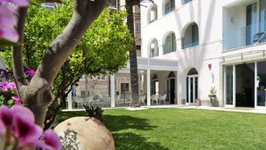 A courtyard with a lawn, white tables, and chairs, surrounded by buildings with arched windows and a large planter. - Hotel Il Piccolo Giardino (Taormina)