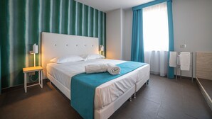 A hotel room with a large bed, a bedside table with a lamp, a window with curtains, and a bathroom area with a towel rack. - Hotel Il Piccolo Giardino (Taormina)