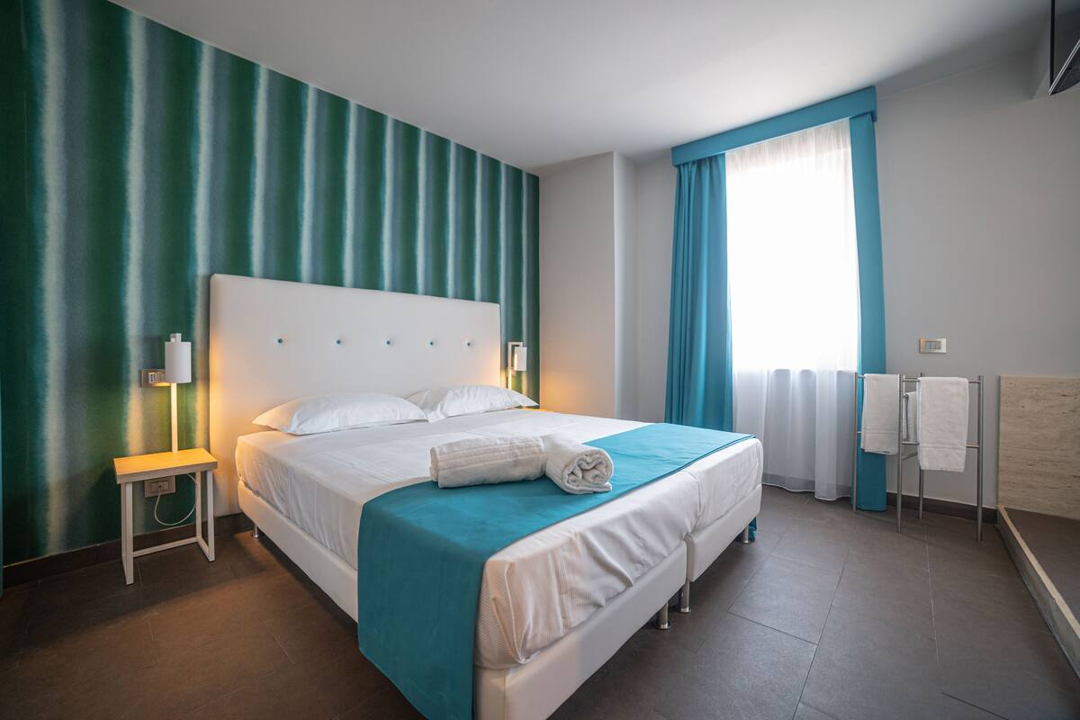 Une chambre d’hôtel dotée d’un grand lit, d’une table de chevet avec une lampe, d’une fenêtre avec des rideaux et d’une salle de bain comprenant un porte-serviettes.