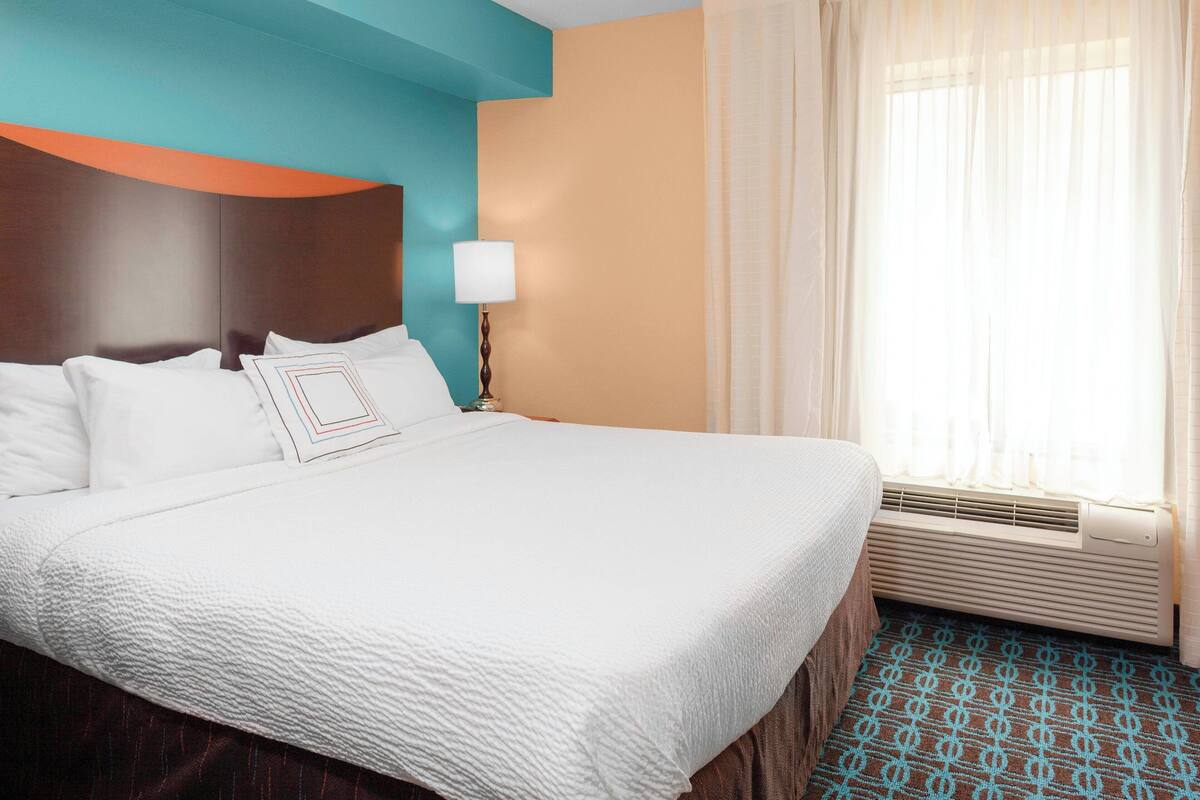 A hotel room with a large bed, a bedside table with a lamp, a window with curtains, and a wall with a teal and orange design.