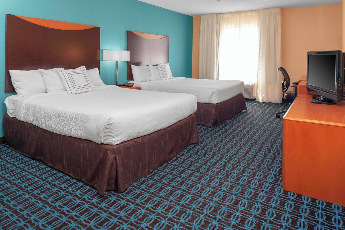 A hotel room with two beds, a desk with a television, and a patterned carpet.