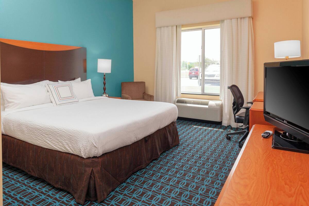 A hotel room with a large bed, a desk with a television, a chair, a window with curtains, and an air conditioning unit.