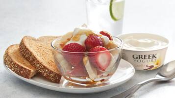 Un desayuno con un bol de vidrio con yogur, fresas y plátanos, acompañado de tostadas y un vaso de agua.