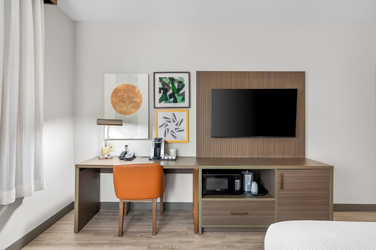 Habitación de hotel con escritorio de madera, una silla naranja, televisor de pantalla plana, obras de arte y un microondas.