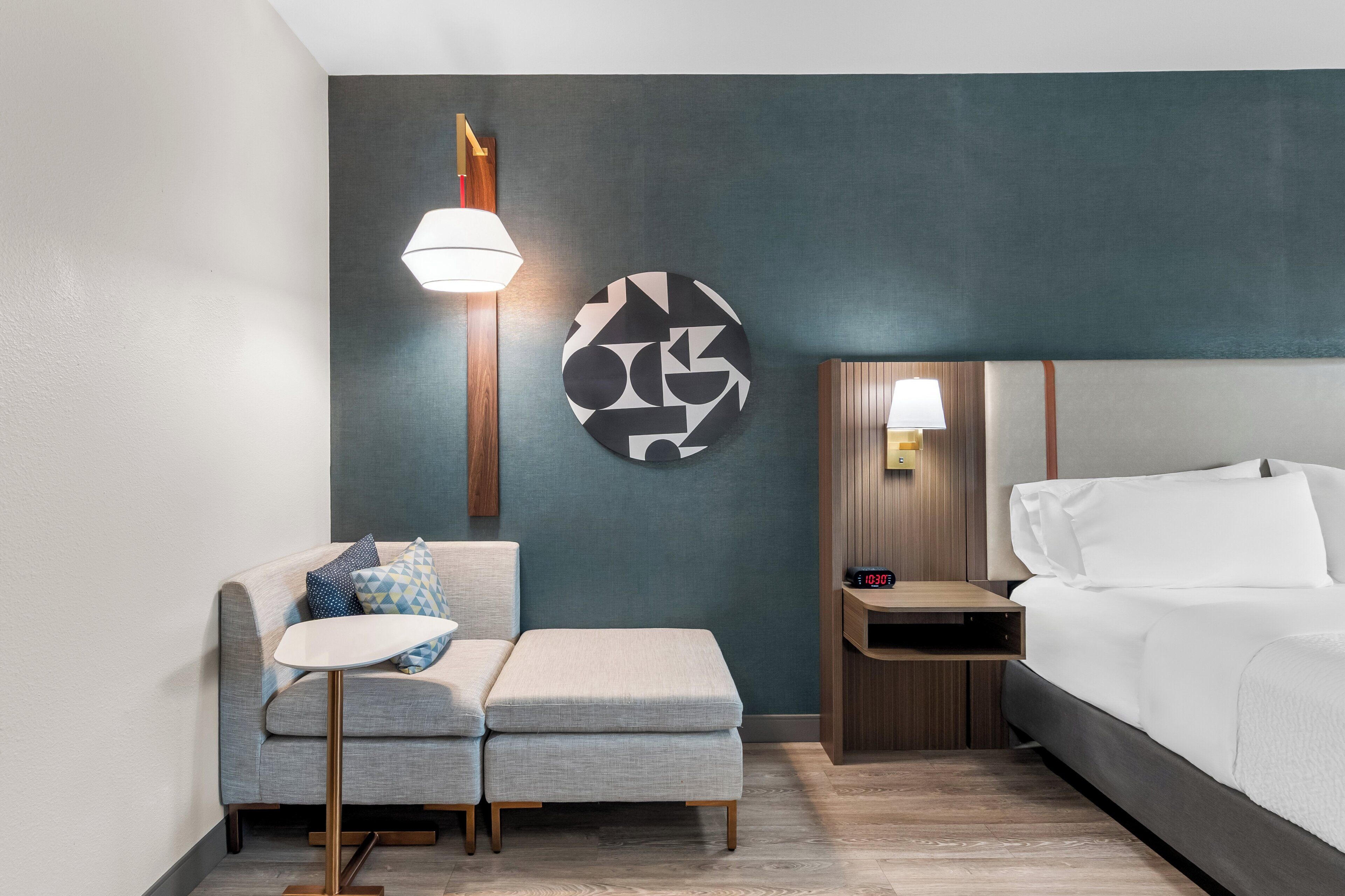 A modern hotel room with a bed, a small sofa, a side table, a wall-mounted lamp, and a clock.