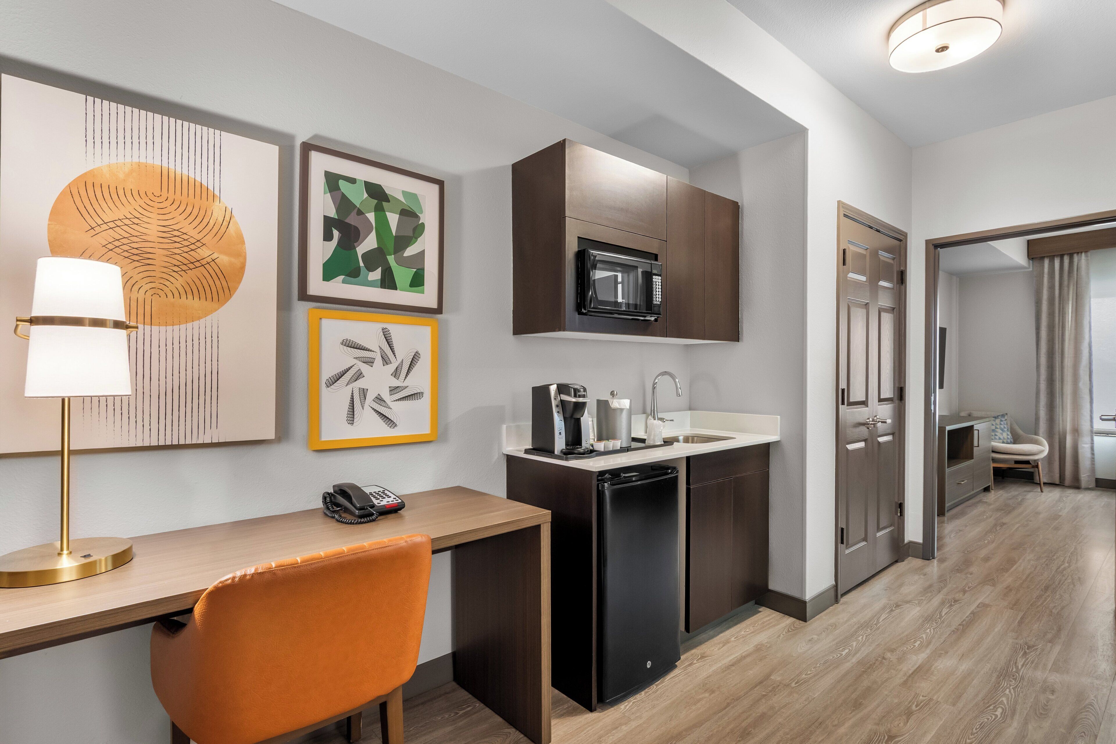 A compact kitchen with a microwave, coffee maker, and dishwasher. There is a desk with a lamp and a phone. The room has a modern design with artwork on the walls.