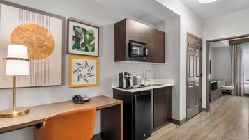 A compact kitchen with a microwave, coffee maker, and dishwasher. There is a desk with a lamp and a phone. The room has a modern design with artwork on the walls.