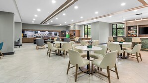A modern restaurant with round tables and chairs, a bar area, and a kitchen in the background. - Holiday Inn Oklahoma City Airport by IHG (Oklahoma City)