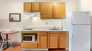 A compact kitchen with wooden cabinets, a white refrigerator, a microwave, and a small dining area with a chair and table. - Extended Stay America Select Suites - Cincinnati - Florence - Airport (Florence)