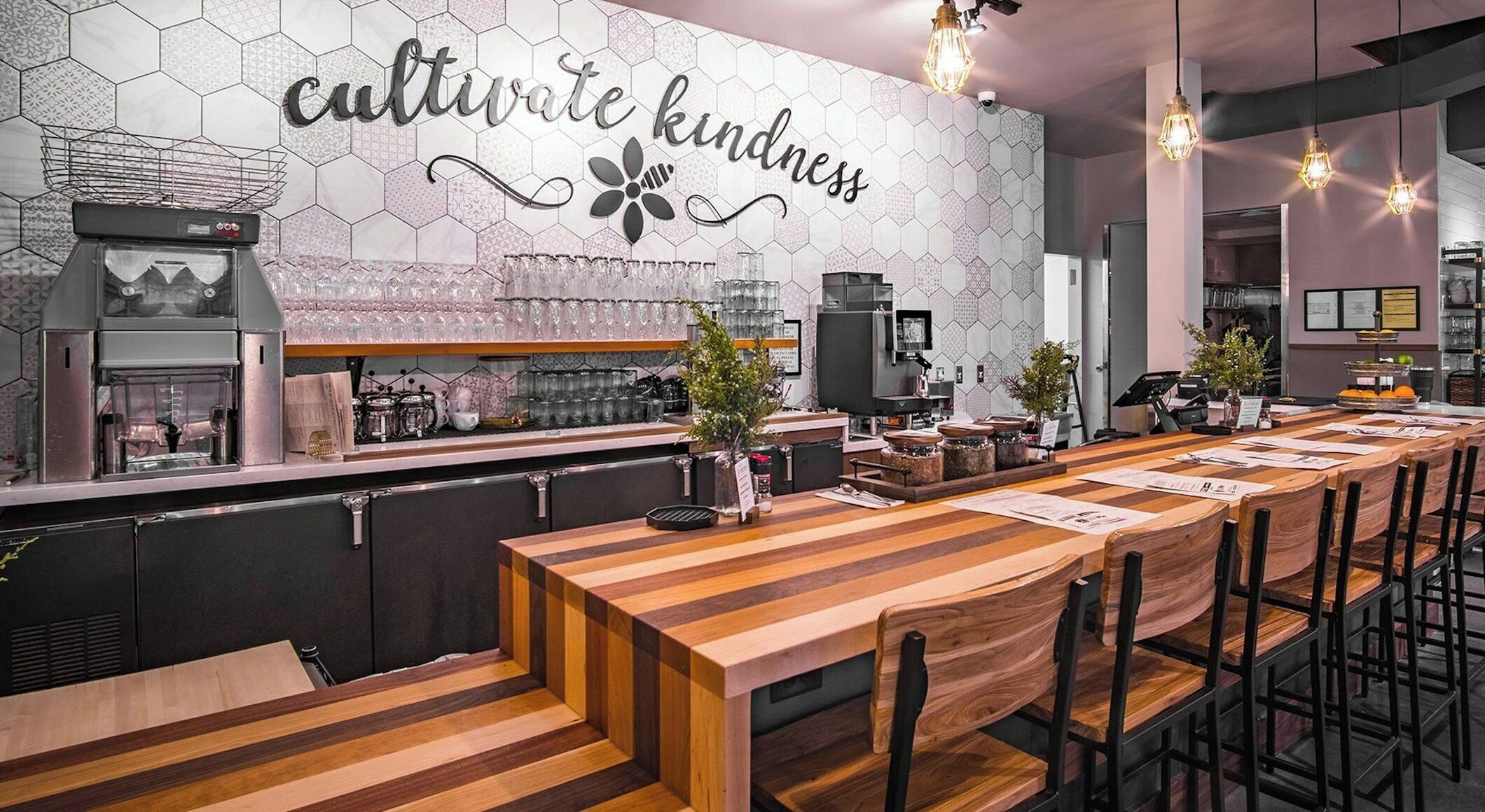Interior de restaurante con una larga mesa de madera, sillas, zona de bar y pared con azulejos hexagonales y un letrero que dice 'cultivate kindness'.
