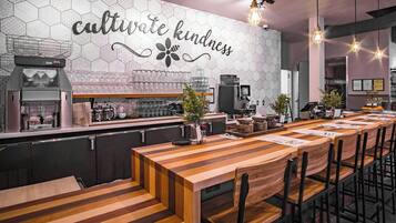 Interior de restaurante con una larga mesa de madera, sillas, zona de bar y pared con azulejos hexagonales y un letrero que dice 'cultivate kindness'.