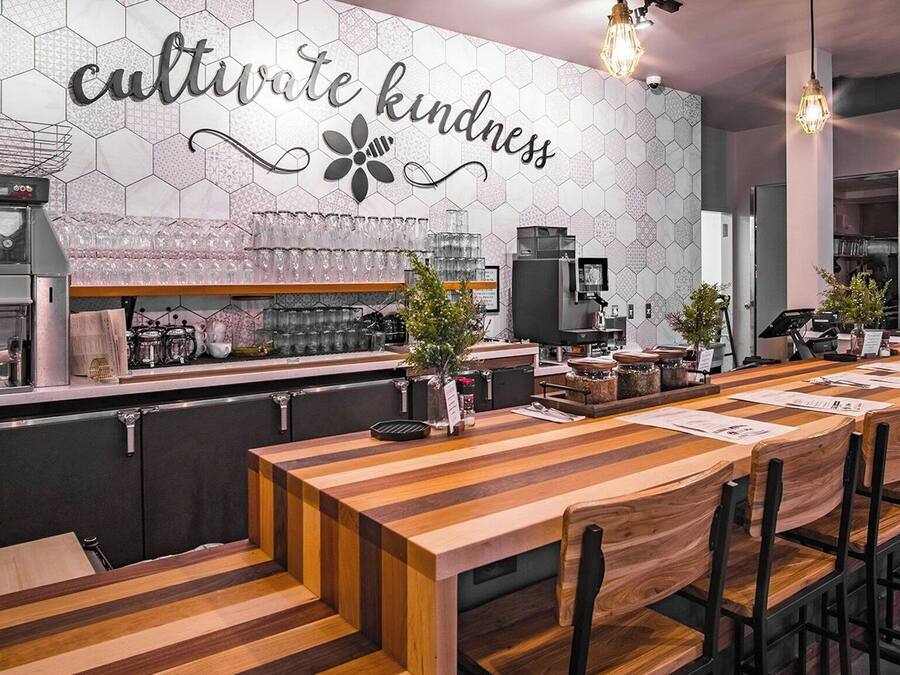 Interior de restaurante con una larga mesa de madera, sillas, zona de bar y pared con azulejos hexagonales y un letrero que dice 'cultivate kindness'.