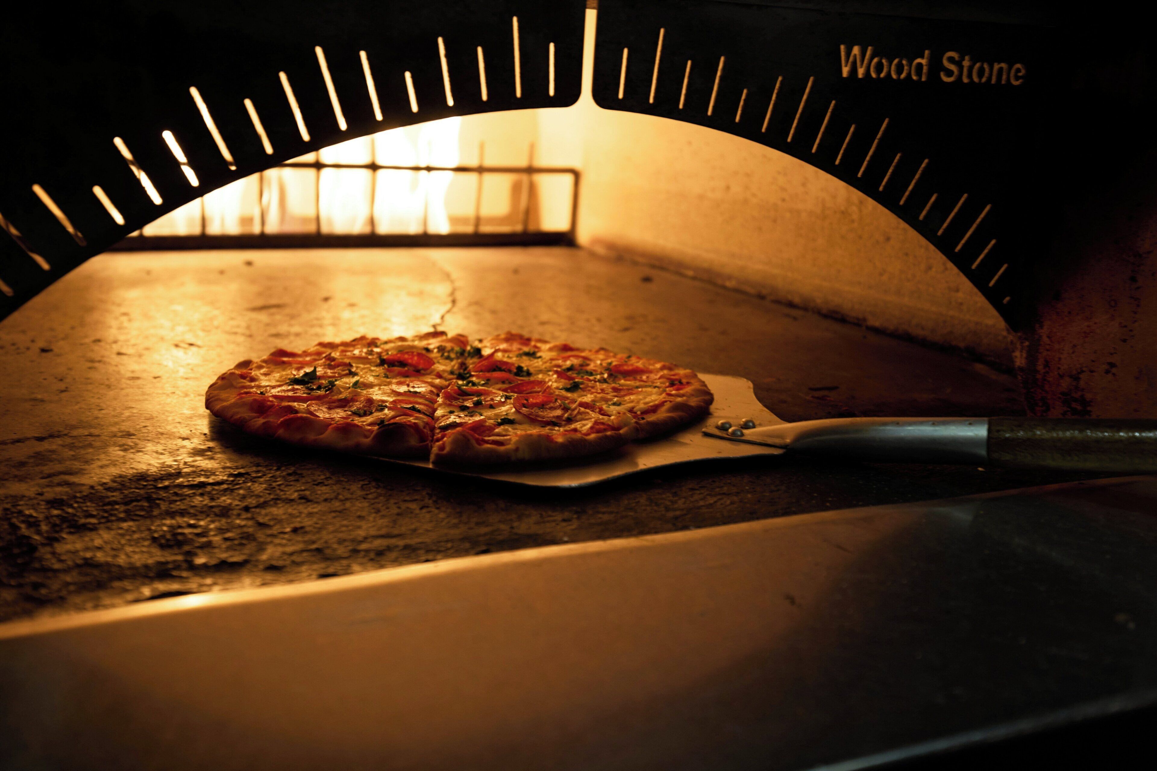 A pizza inside a wood-fired oven with a pizza peel.