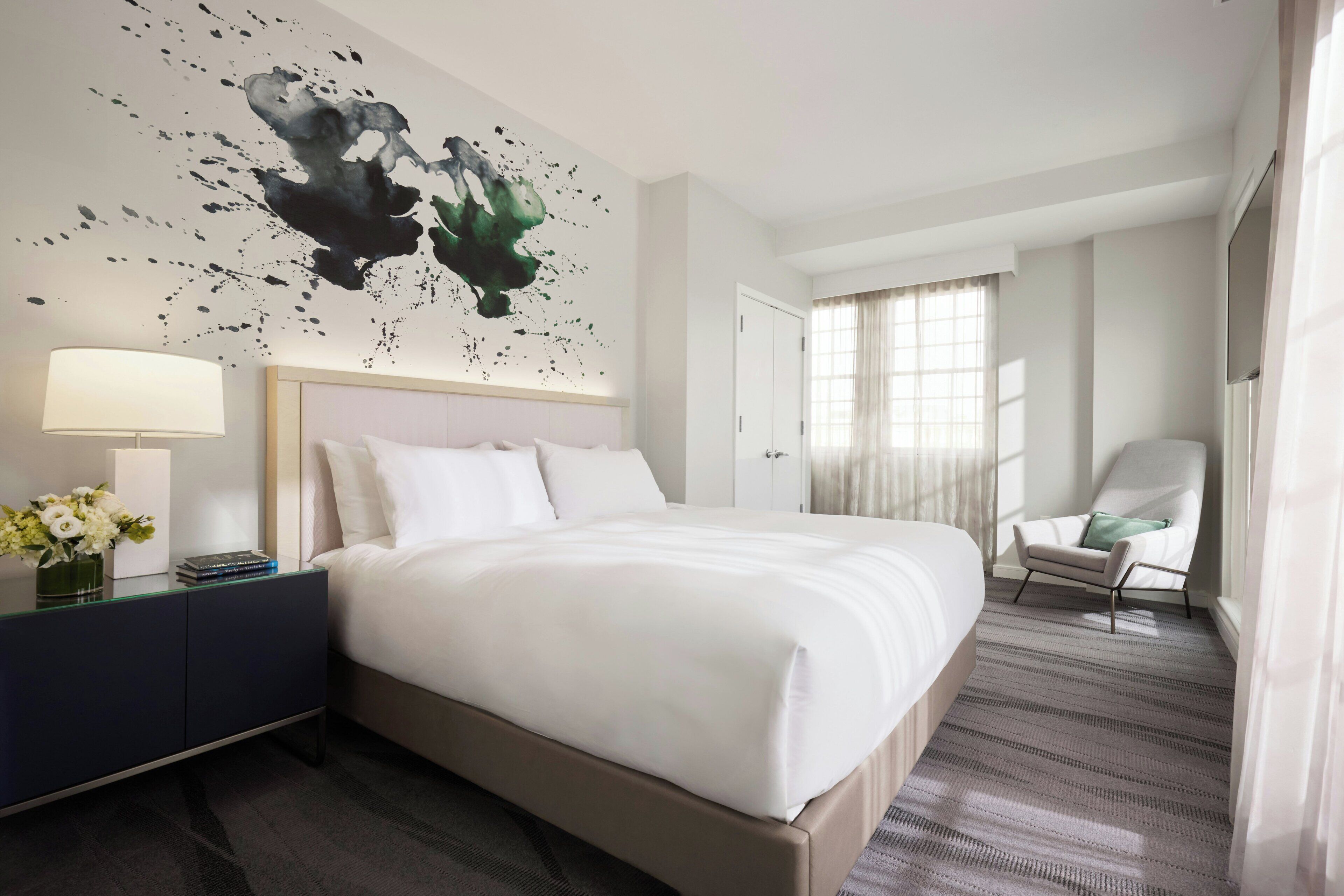 Habitación de hotel con una cama grande, una mesita de noche con lámpara, una silla y un cuadro en la pared.