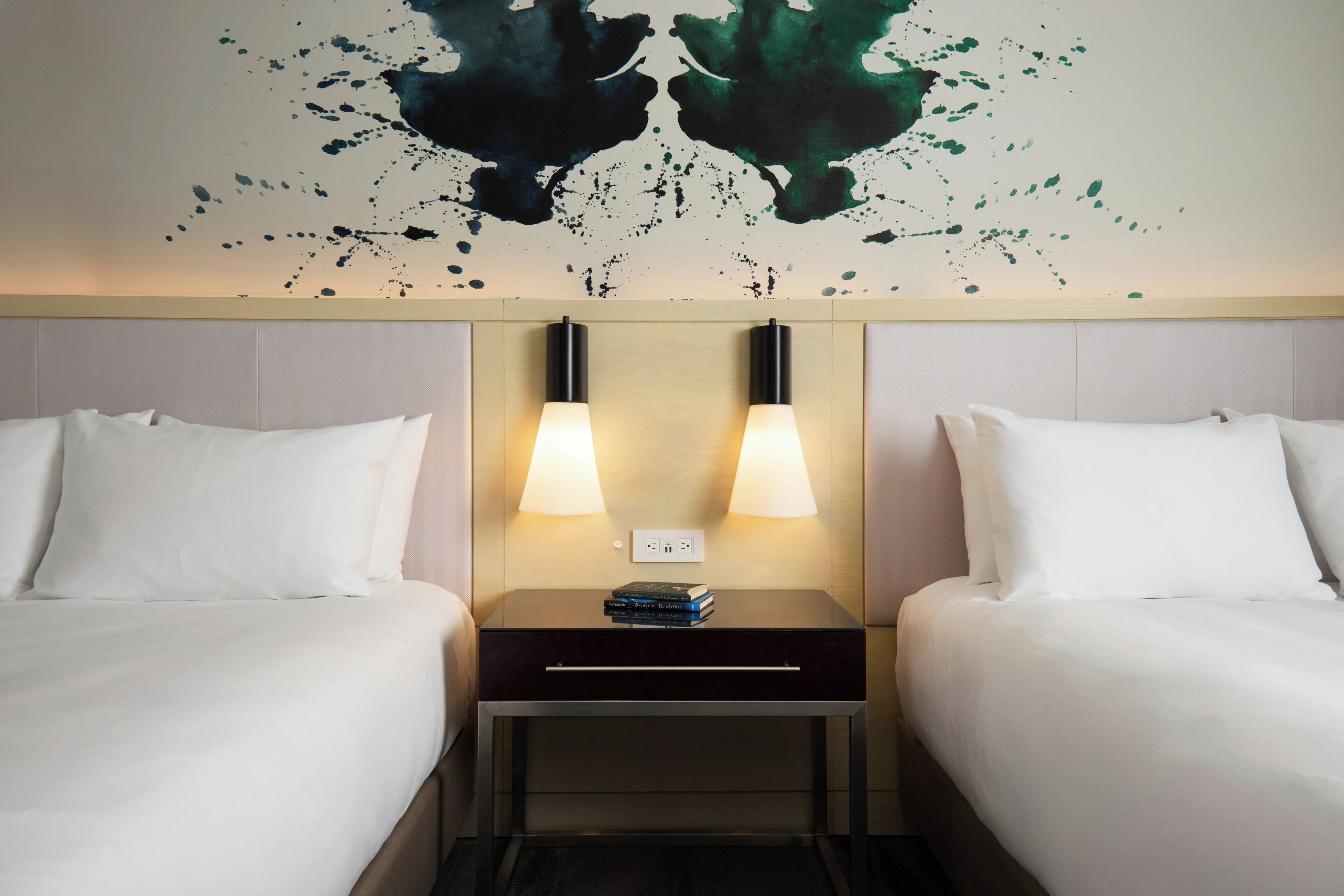 Quarto de hotel com duas camas, mesa de cabeceira com duas luminárias e uma obra de arte na parede acima das camas.