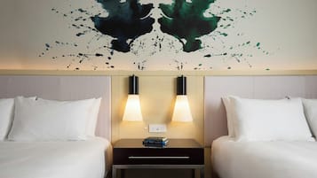 Quarto de hotel com duas camas, mesa de cabeceira com duas luminárias e uma obra de arte na parede acima das camas.