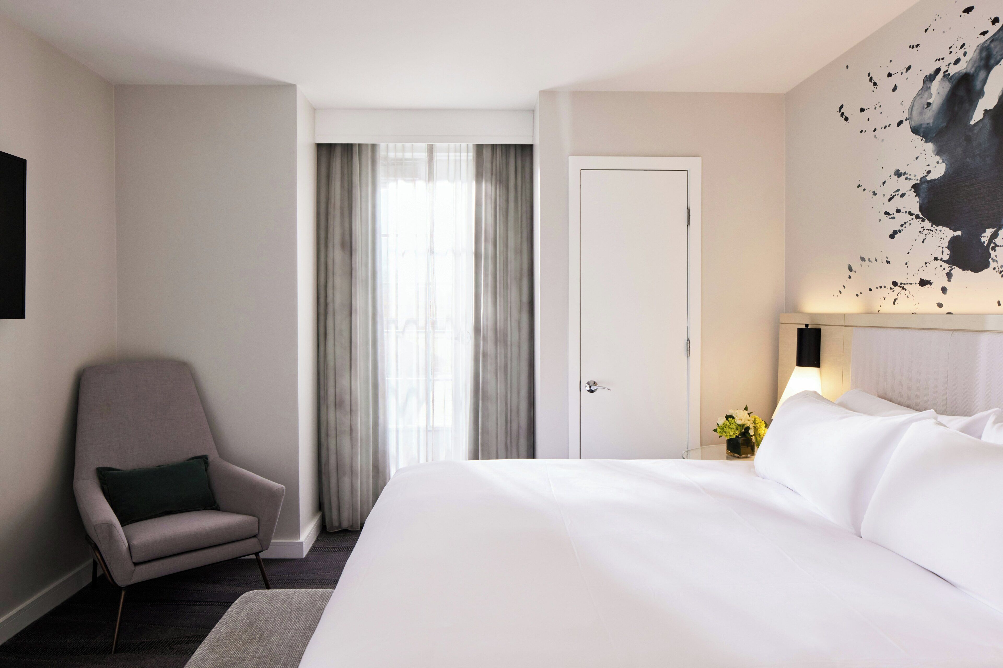 A hotel room with a bed, a chair, a door, and a wall with a painting.