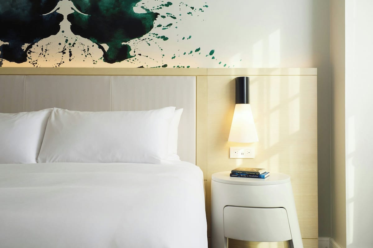 Quarto de hotel com uma cama, uma luminária na parede e uma mesinha com livros.