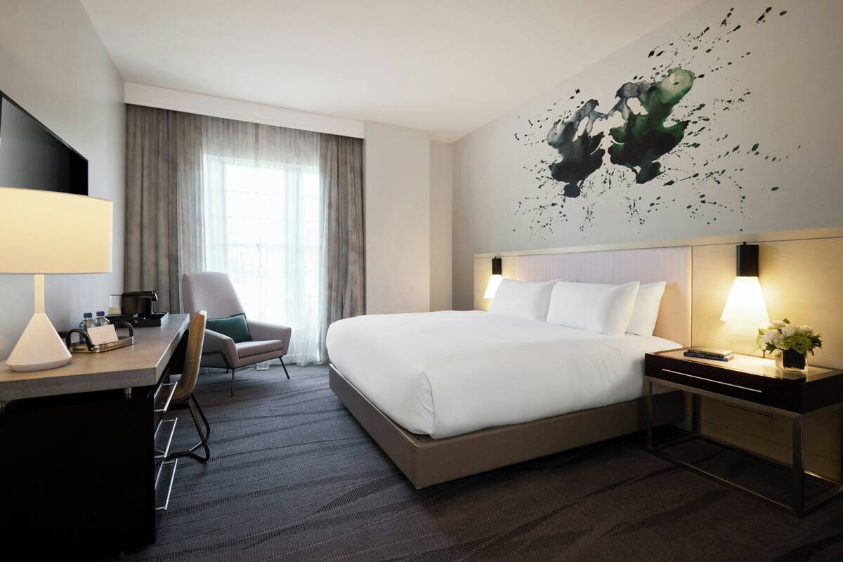 Quarto de hotel com cama, escrivaninha, cadeira e uma parede com um quadro.