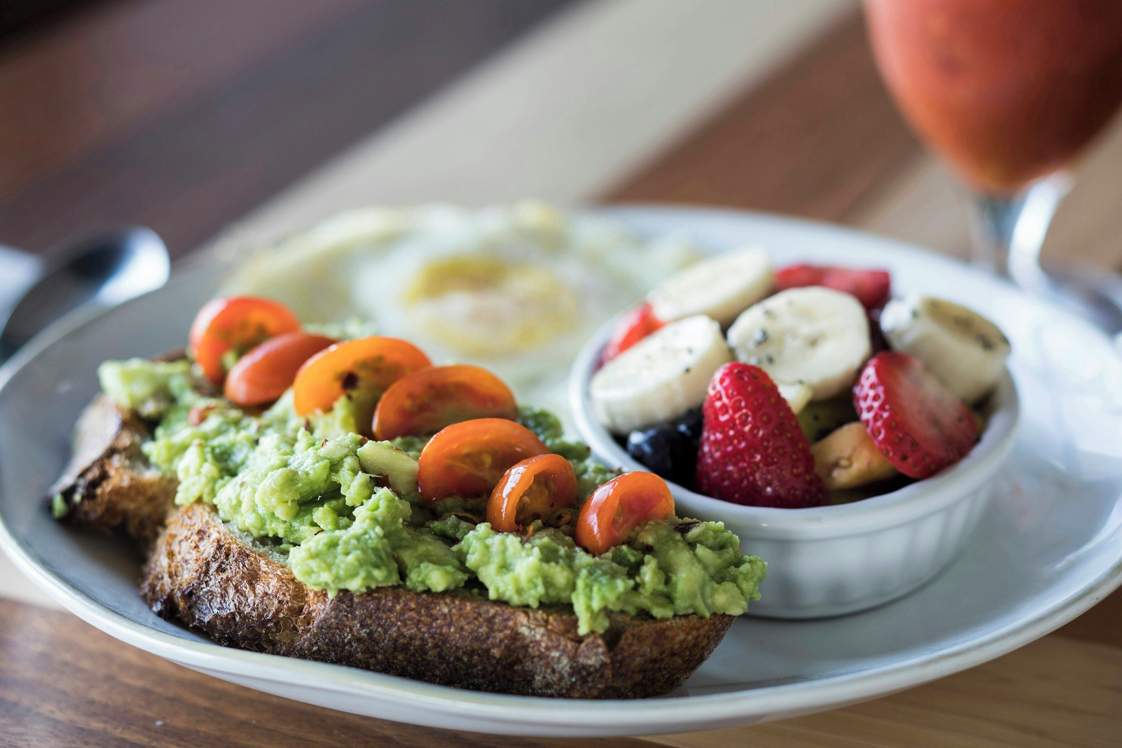 A plate with toast topped with guacamole and tomatoes, a bowl of mixed berries, and a glass of a red beverage.